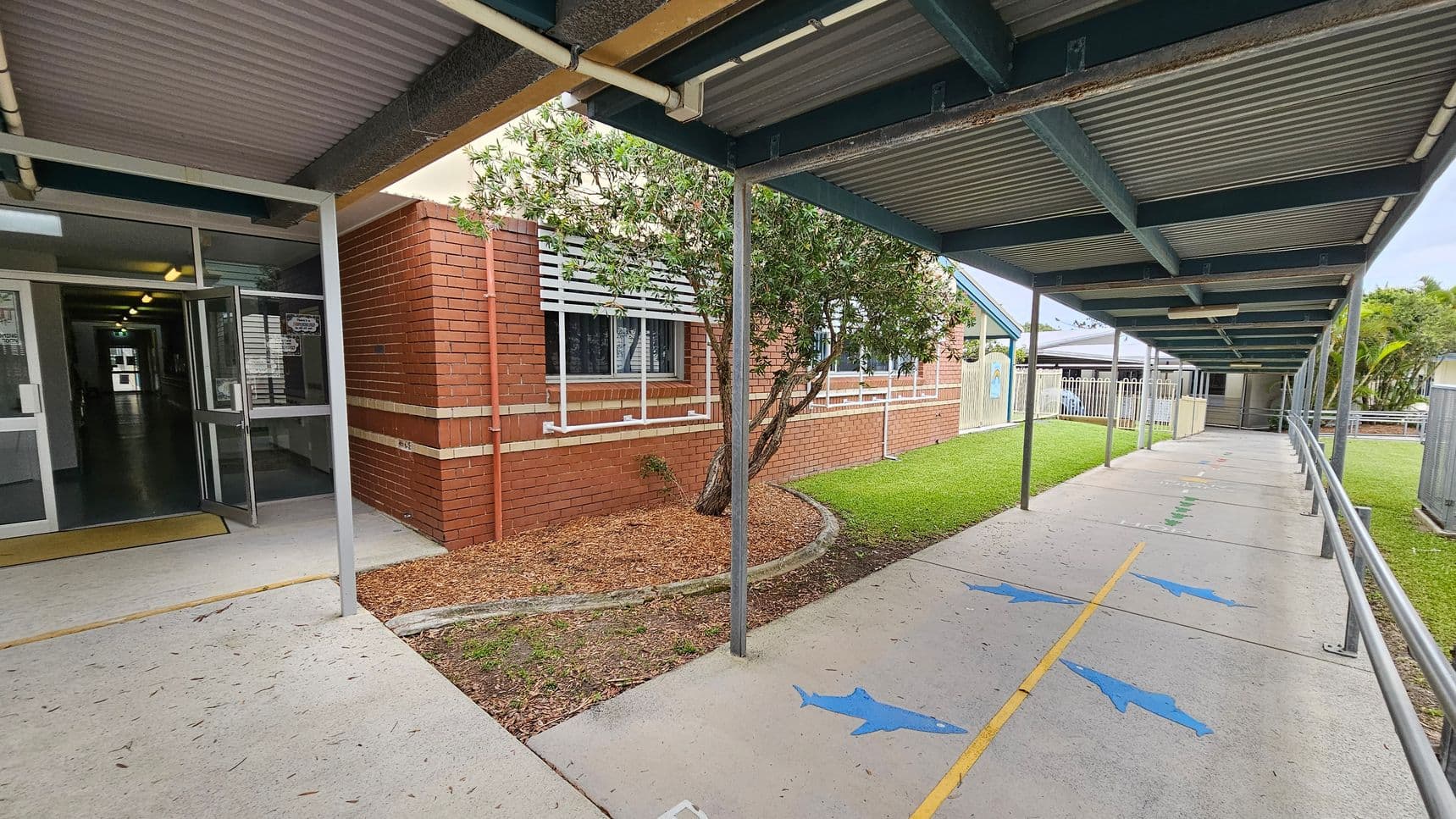 A covered walkway with a corrugated metal roof extends to the right, featuring a concrete path marked with blue dolphin shapes and a yellow line. On the left, a brick building with white window shutters and a small tree in a mulched area is visible. The entrance to the building has glass doors, leading to a lit interior hallway. The grassy area runs alongside the path, bordered by a fence in the background.