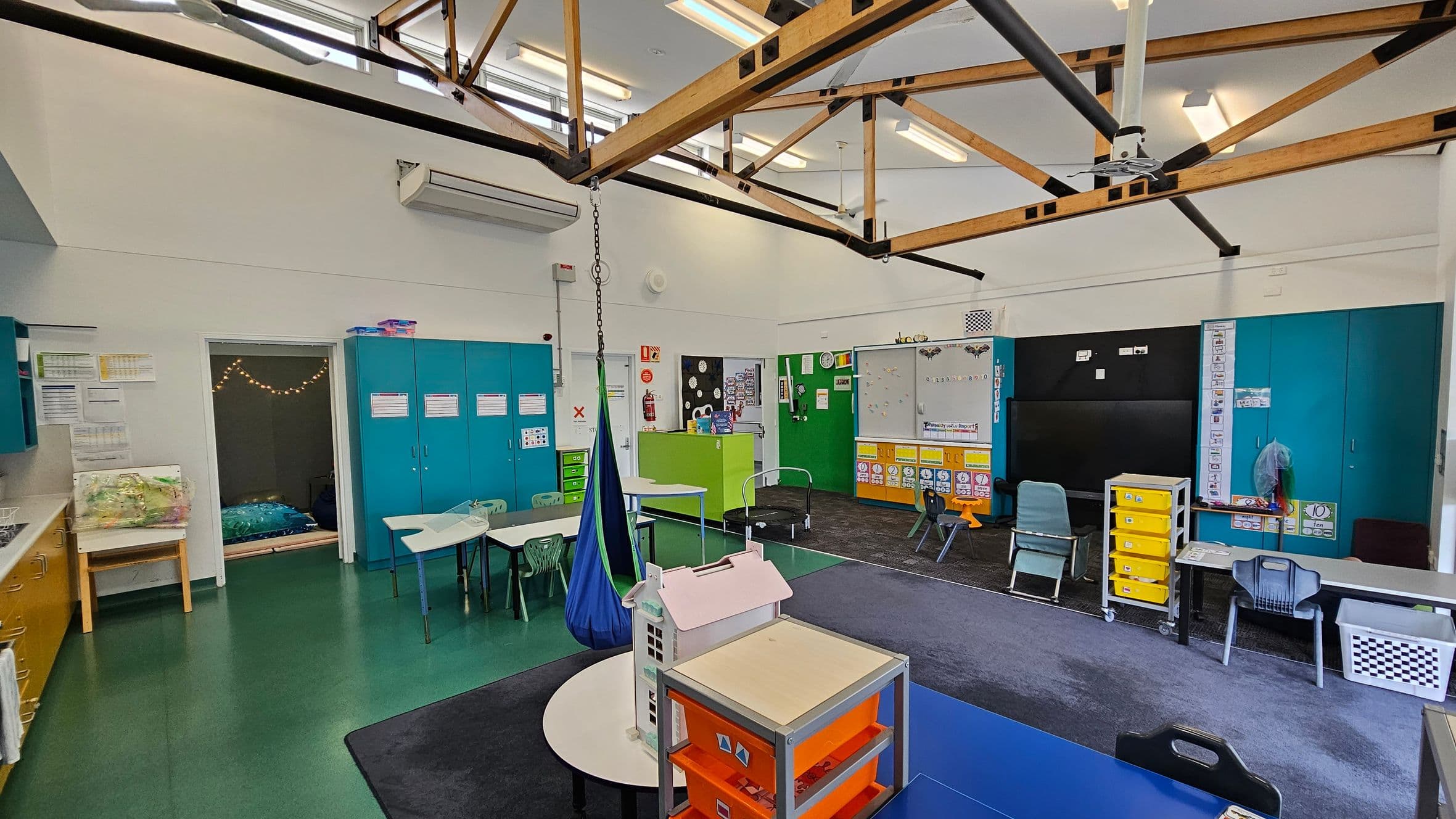 Classroom with green flooring and a high ceiling featuring exposed wooden beams. On the left, there are blue cabinets and a small room with string lights. In the centre, tables and chairs are arranged, with a blue hanging chair. The right side has a blackboard, colourful storage units, and more chairs. A small playhouse and a trolley with yellow drawers are in the foreground. The walls are decorated with educational materials.