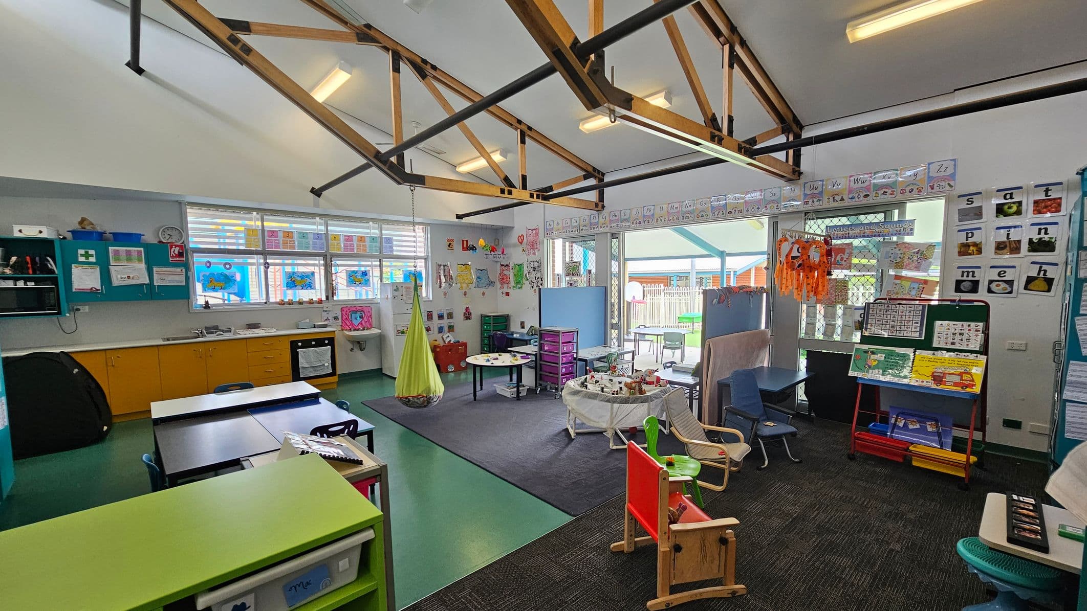 Classroom with a high ceiling featuring exposed beams and fluorescent lights. On the left, there are yellow cabinets and a sink beneath windows decorated with colourful artwork. In the centre, a small round table and a hanging green chair are on a carpeted area. The right side has a display board with educational materials and a variety of chairs. The walls are adorned with alphabet cards and children's artwork. The floor is a mix of green and dark carpet.