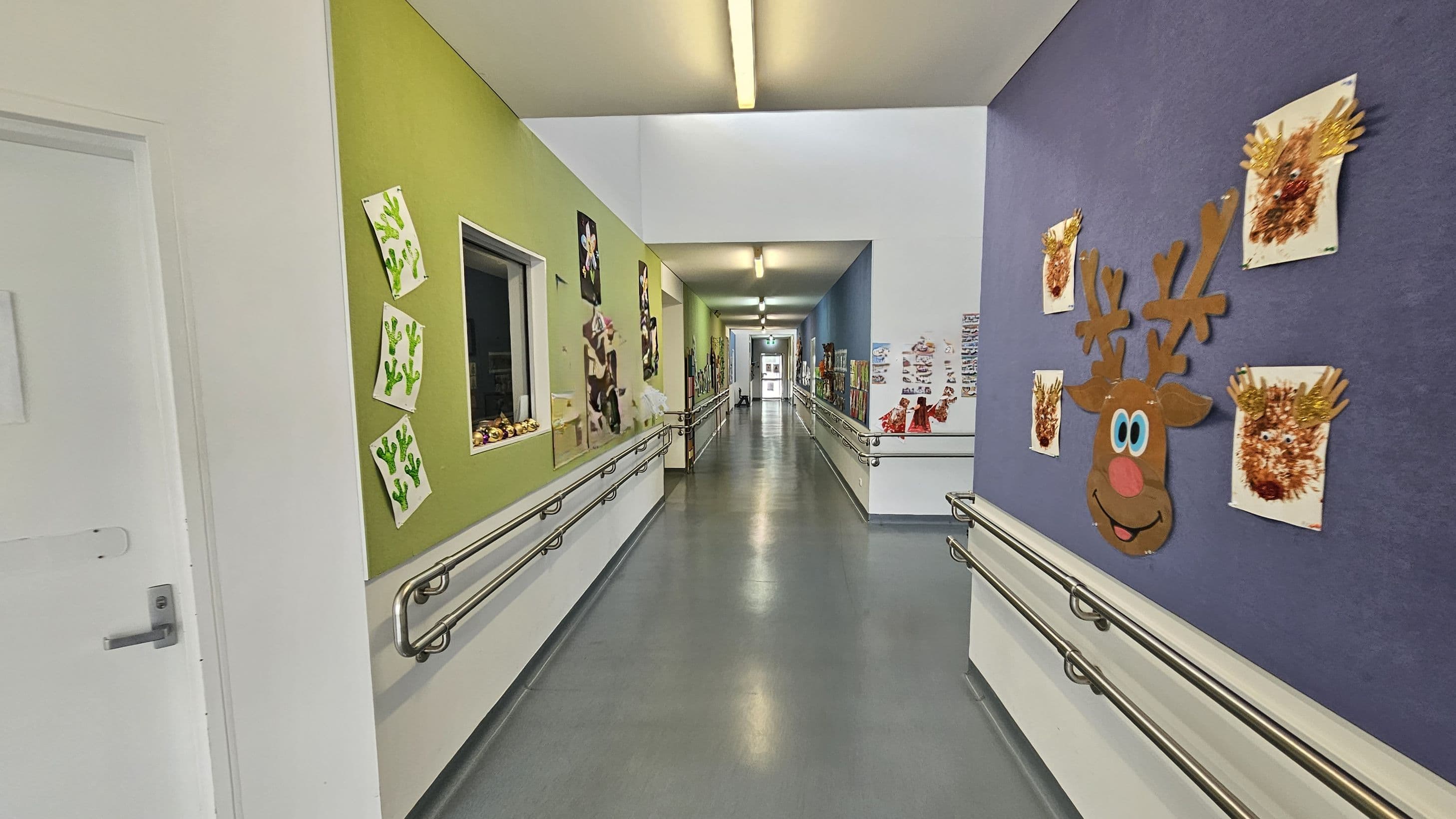 A corridor with colourful walls decorated with children's artwork. On the left, a green wall displays cactus drawings and other art pieces. On the right, a purple wall features a large reindeer face and handprint art. The floor is grey and smooth, with metal handrails on both sides. The ceiling is white with fluorescent lights. The corridor extends into the distance, with more artwork visible along the walls.