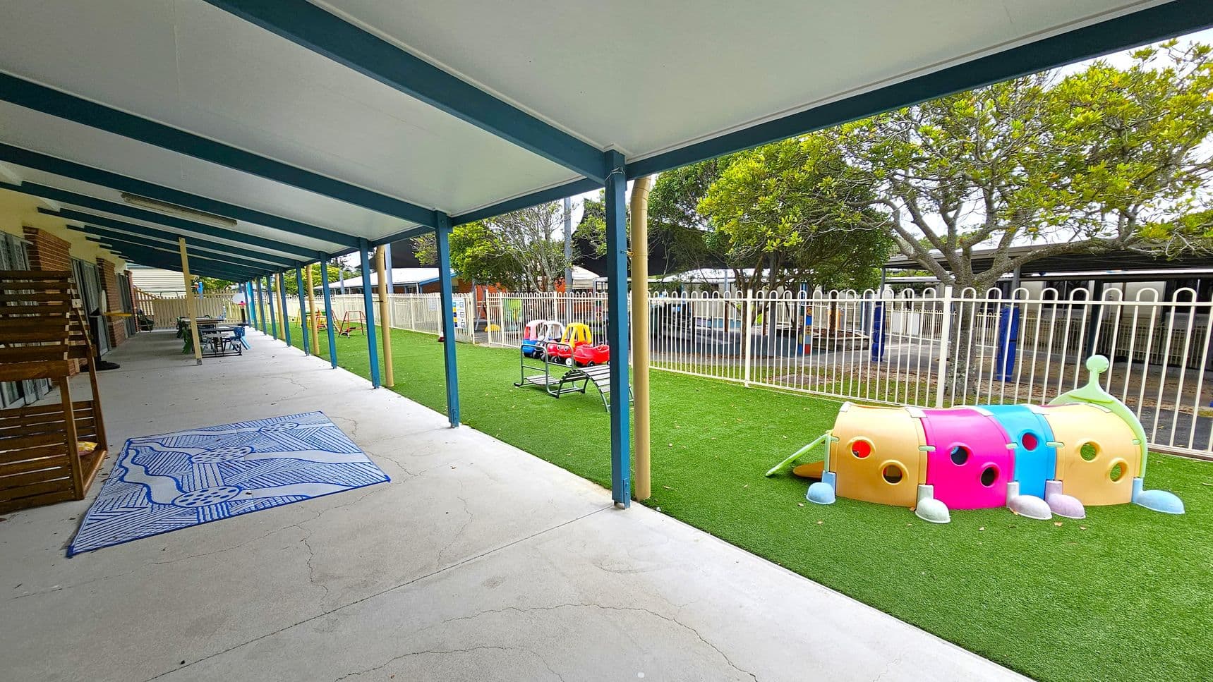 A covered outdoor area with a concrete floor and a blue and white patterned mat on the left. Wooden furniture and tables are arranged along the wall. On the right, a grassy area features colourful play equipment, including a caterpillar tunnel and toy cars. A white fence separates the play area from trees and additional structures in the background. The ceiling is supported by blue beams.