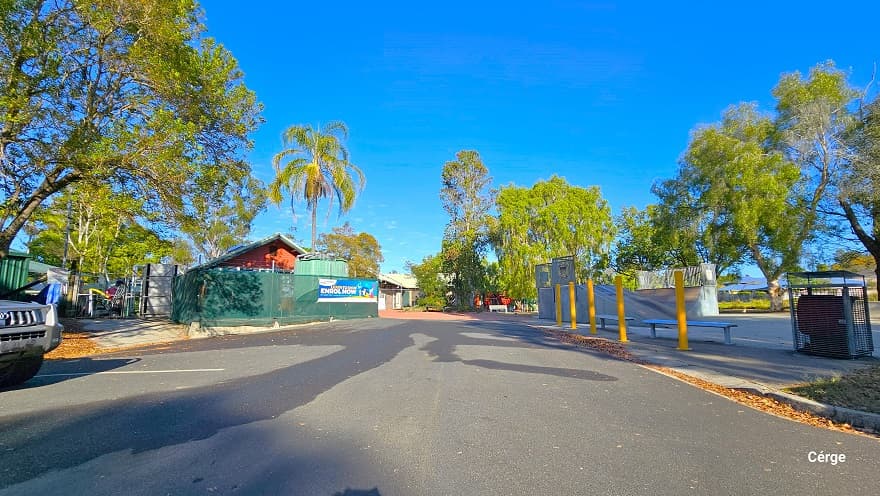 Entryway from the carpark leading to the entrance of Dayboro Pool & Gym. Tall yellow bollards marked the boundary of the skate park on the right side. A large grey metal garbage bin with a canopy cover is placed before two benches at the back of the bollards. Covering the perimeter of the facility is a green cloth on the left, while on the right is white. Tall trees with great foliage offer shade to the surrounding area.