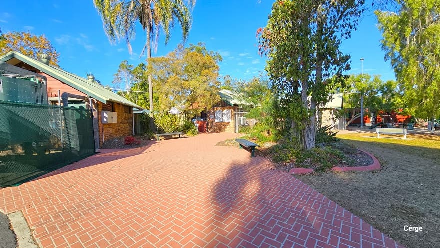 Red brick pathway in Dayboro Pool & Gym. On both edges of the pathway are black benches. The property has low-set buildings with brown tile walls, red wooden front walls, and white roofing structures with round roof vent fans. A green chain link fence encloses the front of the first building with a large grey water tank.