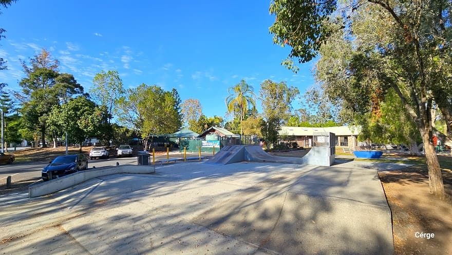 Skate Park in Dayboro has a large open flat space in the middle with an angle ledge on the left and a flat launch ramp on one end. Further to the left are car park spaces separated by white lines. The perimeter of Dayboro Pool and Gym is surrounded by tall trees with a large canopy cover.