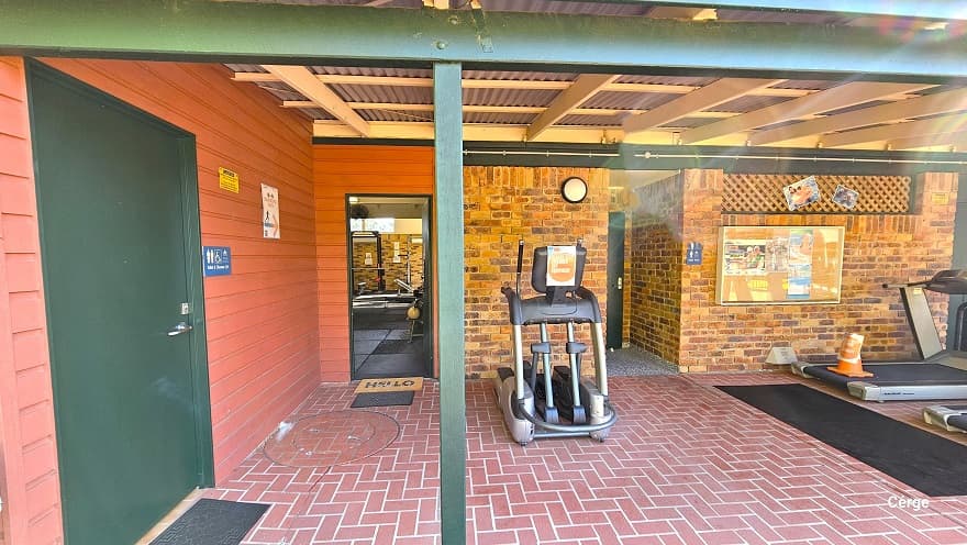 The 1970-style brick building with red corrugated walls and roofing has a red brick floor style. The building has green metal frames as support. Within the red corrugated wall is the accessible bathroom, which can be distinguished by the green door, a black mat, and a blue Unisex Toilet label. On the brick wall to the right is a painting and two photos. Two elliptical trainers are available in this area.
