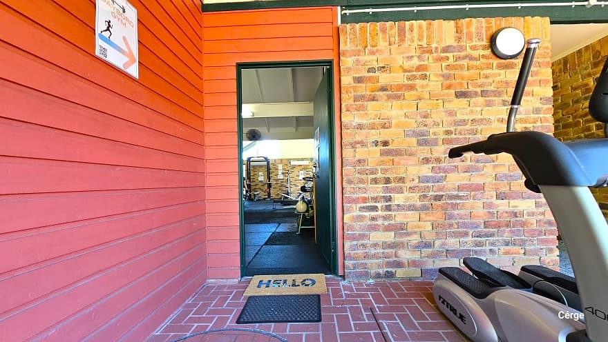 Entryway to Dayboro Gym. The gym can be identified by the red-to-orange brick wall and red corrugated wall and a directional white label placed within this wall that details the Dayboro Gym with an arrow pointing to the door. There is a brown floor mat with a Hello detail on it and a black mat for dirt removal before entering the amenity. Found on the right is cardio equipment called an Elliptical trainer.