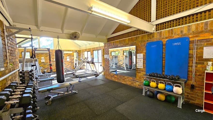Inside the Dayboro Gym are black dumbbells, a row of colorful kettlebells on a rack, and other cardio endurance equipment. The room has a wooden netted-like design on the upper right wall that has white metal frames. Behind the rack for dumbbells and kettlebells are blue exercise mats and grey ropes. Next to it is a mirror that reflects other gym equipment inside. Further to the back, on the right wall, are glass windows that allow natural light to filter through, illuminating the area inside.