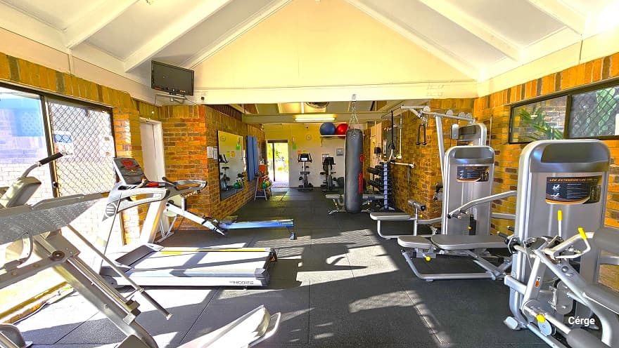 The image shows the back portion of the inside of Dayboro Gym. Mounted on the left wall is a small television above another white door. The glass windows reflect the silhouette of the chain-linked fence outside the building. On the upper left of the white wall next to the entrance door, there are blue and red yoga balls placed on a white rectangular frame. Two small glass windows can be found on the upper part of the right brick wall.