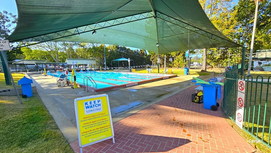 Closer of the six-lane, 25m lap outdoor pool. A yellow sign board that reminds adults to keep watch for children of different age brackets and what lifeguards do can be found on the edge of the red brick pathway that extends to the right. Within this pathway is a grey metal bench, and a blue container with black wheels that contains lime green and blue plastic tubes. There is an aquatic pool and a yellow pool hoist on the left pool deck.
