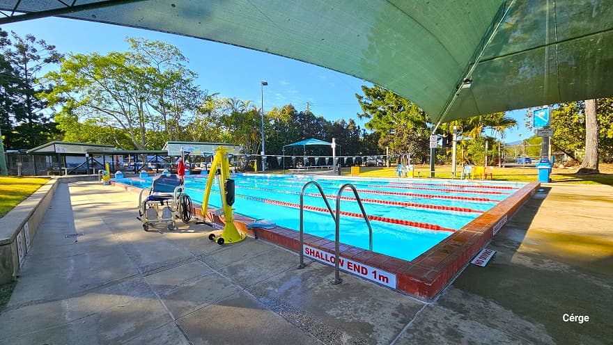 A 25m, 6-lane outdoor pool at Dayboro Pool and Gym with partial shading on one end. The pool has red-tiled edges all around with stainless steel pool ladders. A ‘Shallow end 1m’ and ‘No smoking’ sign is visible on the edge of the pool. There is an accessible aquatic wheelchair and a yellow pool hoist on the left pool deck. Each lane is separated by blue, red, and white floating rope lanes. Overhang the pool is strings of grey triangular flags wrapped around a pole on the opposite pool sides.