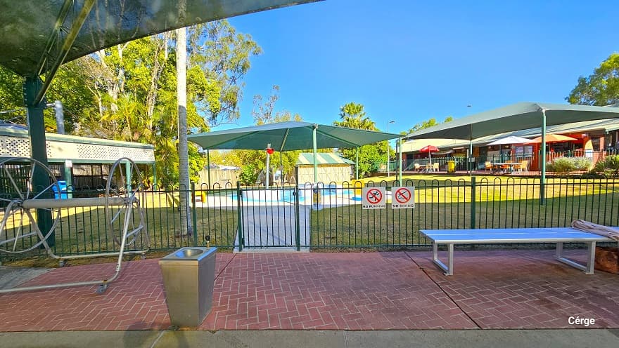 Red brick pathway next to the toddler splash pool enclosed by a green pool fence. Hanging on the green pool fence are the No Running and No Diving white signboards. There is a silver hose reel, a stainless steel wash basin, and a silver metal bench. Further to the left is a long sunshade with an upper side white cover that has several holes as designed.