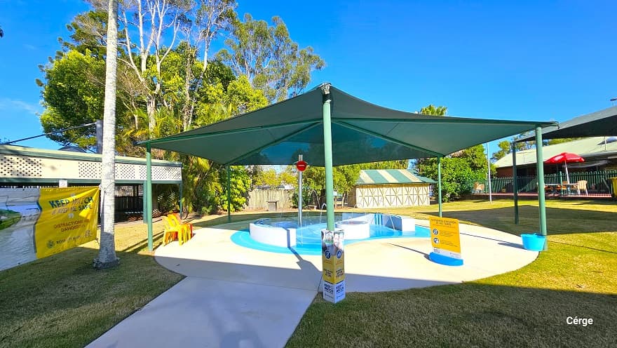 There is a smooth concrete pathway leading to the small toddler splash pool at Dayboro Pool & Gym. The pool is protected by a green sunshade wrapped around green posts. On a silver metal post is a large red lip graphic placed near the top of the post. The splash toddler aquatic activity offers interactive narrow water fountains from the two opposite white curves. There are yellow and red monoblock plastic chairs on the pool deck.