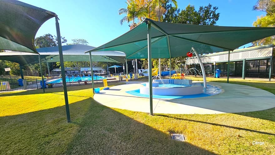 The image shows the side of the small toddler splash pool. The red lip graphic is attached to a red semi-circle on a curvy silver post. The splash toddler aquatic activity offers interactive narrow water fountains from the two opposite white curves. There are yellow and red monoblock plastic chairs on the pool deck. Further to the right is the long sunshade with an upper-side white cover that has several holes. At the back is the 25m, 6-lane outdoor pool.