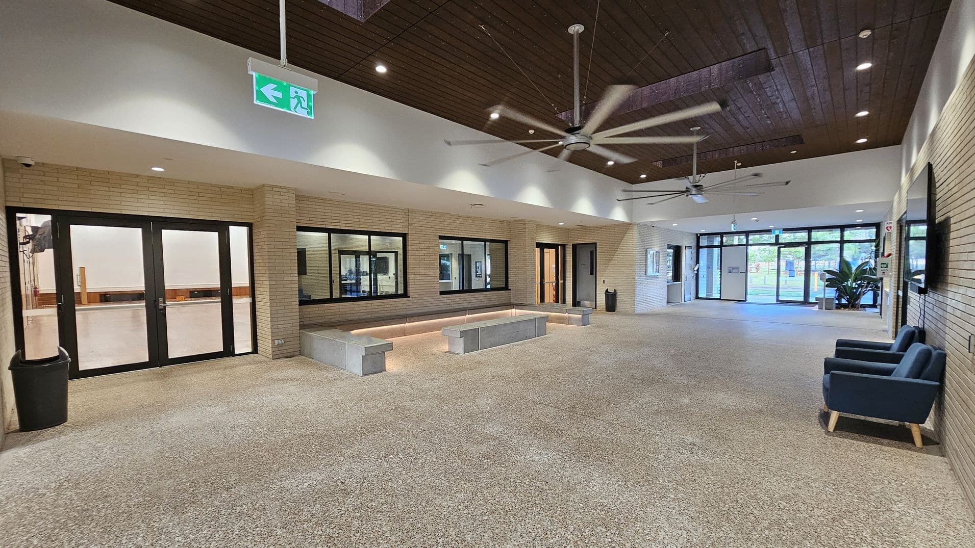 Spacious interior with a terrazzo floor and a wooden ceiling featuring large ceiling fans. On the left, there are black-framed glass doors and windows set in a light brick wall, with a black bin nearby. In the centre, a built-in seating area with concrete benches and soft lighting. On the right, two blue armchairs are placed against the wall, and large glass doors lead outside. An illuminated green exit sign hangs from the ceiling.