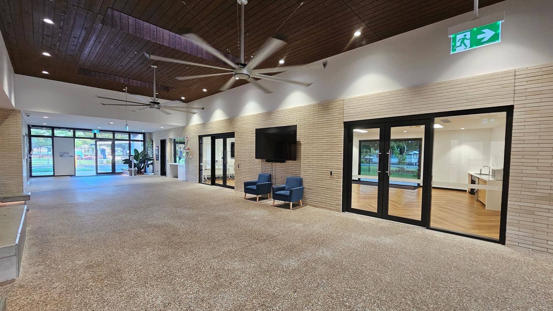 A spacious interior with a polished stone floor and a wooden ceiling featuring recessed lighting and large ceiling fans. On the right, there are glass doors leading to a room with wooden flooring and a kitchenette. Two blue chairs sit beneath a wall-mounted TV. On the left, large windows and doors provide a view of greenery outside. An emergency exit sign is visible above the doors.