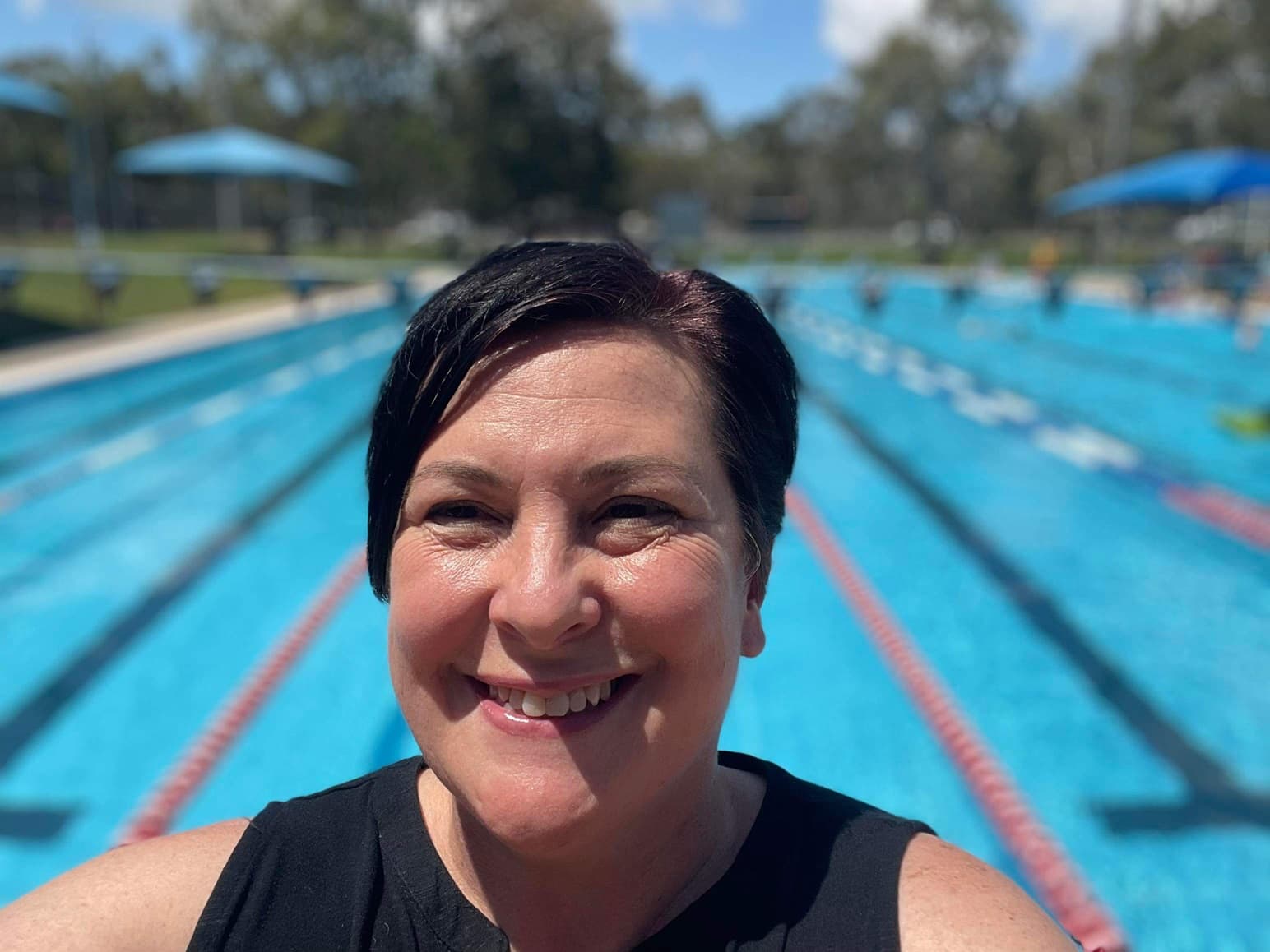 A smiling woman takes a selfie at an outdoor pool with lanes.