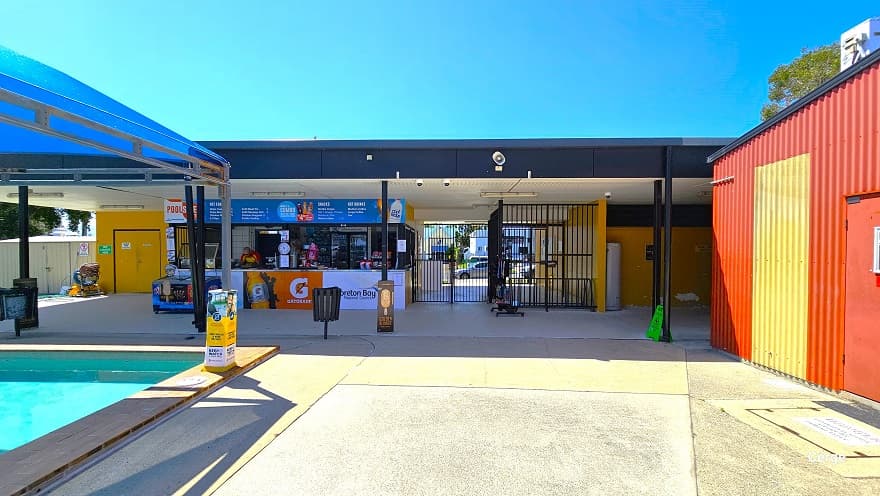 View of the central entry with black iron rod gates and the reception. On the left side of the reception with retail is a small space that leads to the male and female bathrooms, plus an accessible bathroom with a press-button entry. The Cérge app logo holding a QR code is placed behind a blue fridge. A small section of the outdoor pool covered with blue sunshade sails and the red corrugated with yellow accents can be seen on the opposite sides.