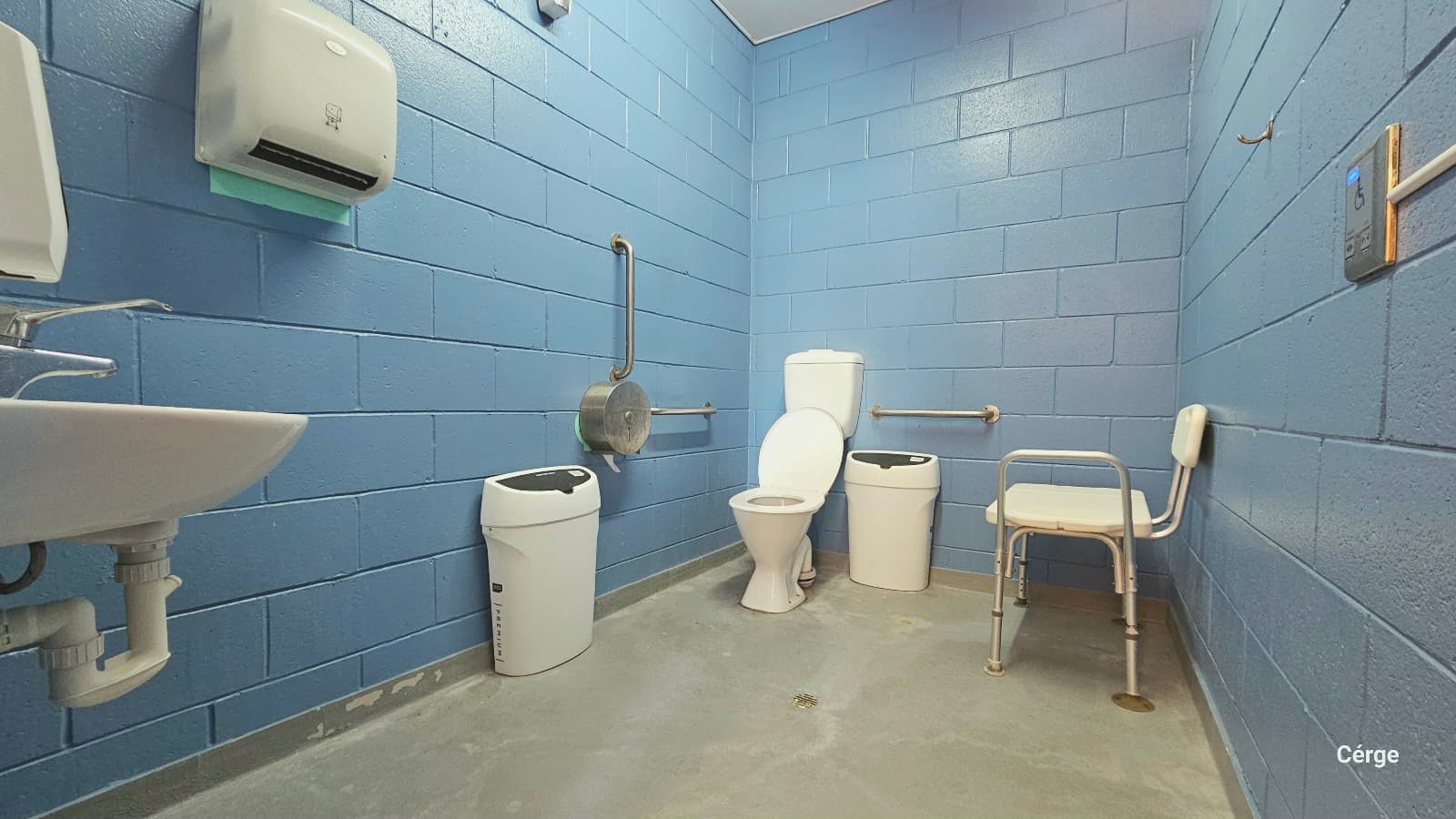 Inside view of the accessible bathroom at Deception Bay Swimming Pool. It has a light blue brick wall, white ceiling, and smooth grey floor. Found near the door are a wash basin, a liquid hand soap dispenser, and a white tissue paper dispenser mounted on the left wall. On the front and side of the toilet are two water softeners. Grab bars mounted on the walls and a white shower and bath seat are available to aid persons with mobility issues.