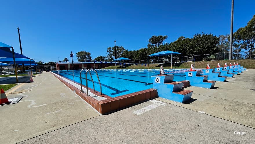 50m, 8-lane outdoor Olympic lap pool. On top of each of the blue 3-tiered stair steps numbered from 1 to 8 is an orange cone. The pool has a dark brown tile pool coping. Each pool lane is separated by blue, white, and red floating rope lanes. Strings of blue triangular flags overhang the pool. On the left side of the pool is a pool ladder with an inverted U handrail. The pool deck is a concreted floor with a rough surface.
