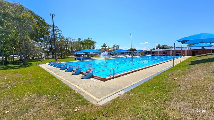 Outdoor 50m 8-lane pool, children’s splash water park, infants shallow pool, and heated learn-to-swim pool, as well as open grass areas for picnics and a large undercover BBQ at Deception Bay Aquatic Centre. The 50m, 8-lane pool has 3-tiered steps numbered from 1 to 8 for diving. On the left and right side of the pool are blue shade sails with seating for families. To the back is a rusty red-colored industrial building with yellow rectangular wall accents and a light blue roof.