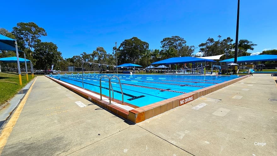 50m, 8-lane outdoor pool has 3-tiered blue steps numbered from 1 to 8 for diving. The pool deck has a noticeable yellow outline close to the border. On the left and right side of the pool are blue shade sails with seating for families. To the back is a rusty red-colored industrial building with yellow rectangular wall accents and a light blue roof. The tall trees offer substantial shading options and the open grass area around the outdoor aquatic area offers open space for an outdoor activity.