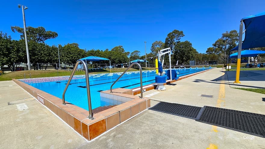 View of the stair access with steel handrails of the 50m, 8 lane Olympic outdoor pool. The image shows that the pool wall is white tile while the pool coping and stairs are a dark brown tile floor. There is a small and large black mat close to the pool stairs. Closer to the pool stairs is a white pool hoist with blue seating to help move persons with mobility disabilities. A No Diving sign can be found on the side of the dark brown pool coping.