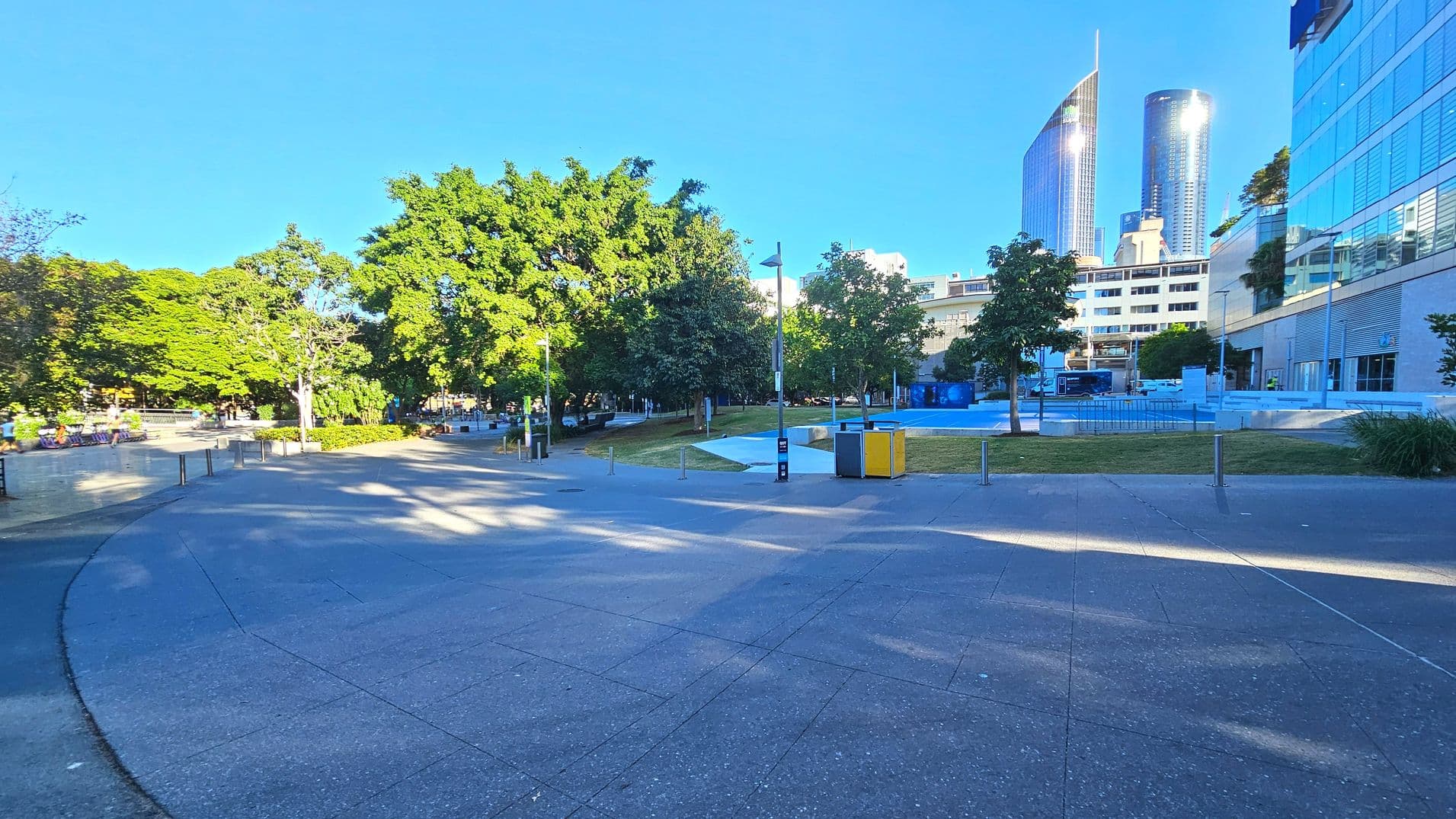 A spacious urban area with a wide, curved concrete path leading through a park-like setting. On the left, lush green trees provide shade over benches and a low wall. To the right, modern glass buildings rise, reflecting light. A small grassy area with scattered trees and a few bollards separates the path from the buildings. A lamppost and a pair of yellow and grey bins are visible near the centre.