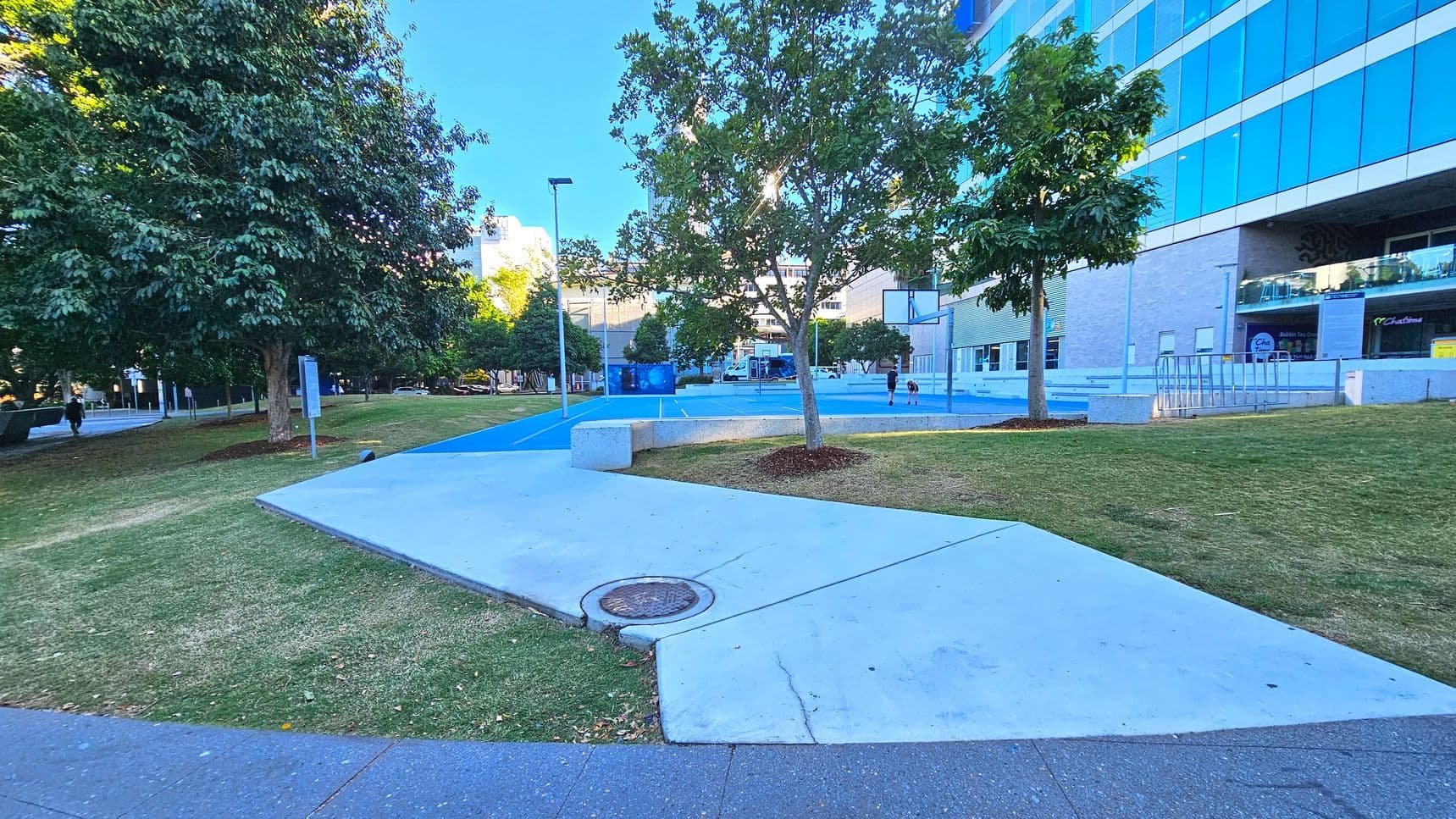 A modern outdoor area featuring a blue basketball court with a hoop, surrounded by grass and concrete paths. A large building with reflective glass windows is on the right, and several trees provide shade. A concrete bench is near the court, and a bike rack is visible against the building. The area is open and spacious, with a mix of natural and urban elements.