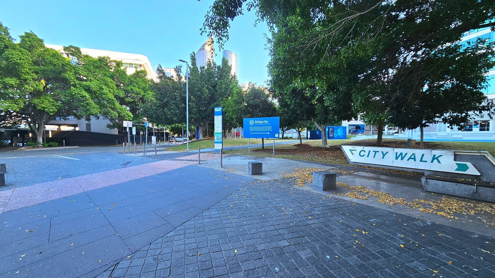 A paved area with a large 'City Walk' sign pointing right is visible. The ground is covered with grey and pink tiles, transitioning to a cobblestone pattern. Several trees with dense foliage line the area, providing shade. A blue sign with white text stands near the centre, and a tall building is partially visible in the background. Metal bollards and a lamppost are also present, indicating a pedestrian-friendly zone.