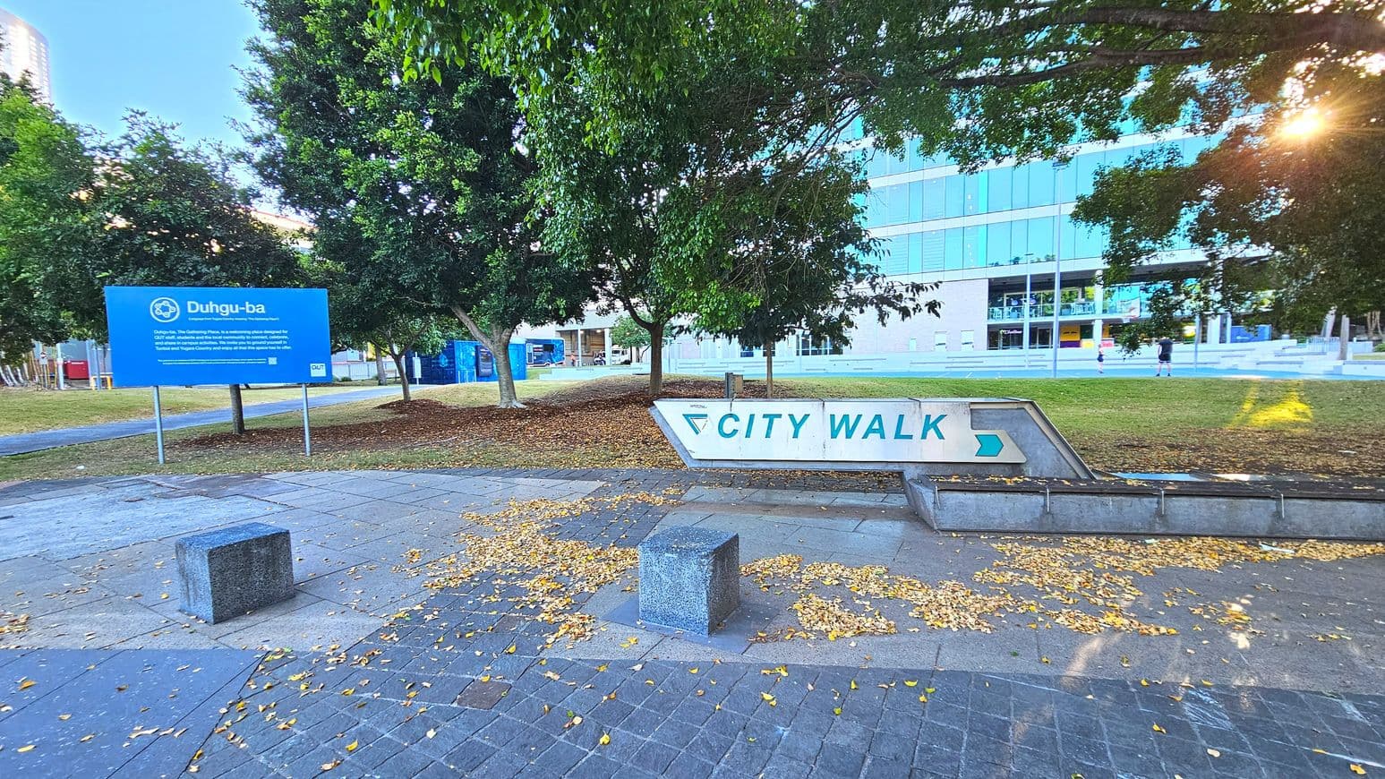 A paved area with a large white sign reading 'CITY WALK' in blue letters and an arrow pointing right. The sign is set on a low concrete structure. Nearby, a blue sign with 'Duhgu-ba' is visible, mounted on metal posts. The area is bordered by trees with scattered fallen leaves on the ground. In the background, a modern glass-fronted building is visible, with steps leading up to it.