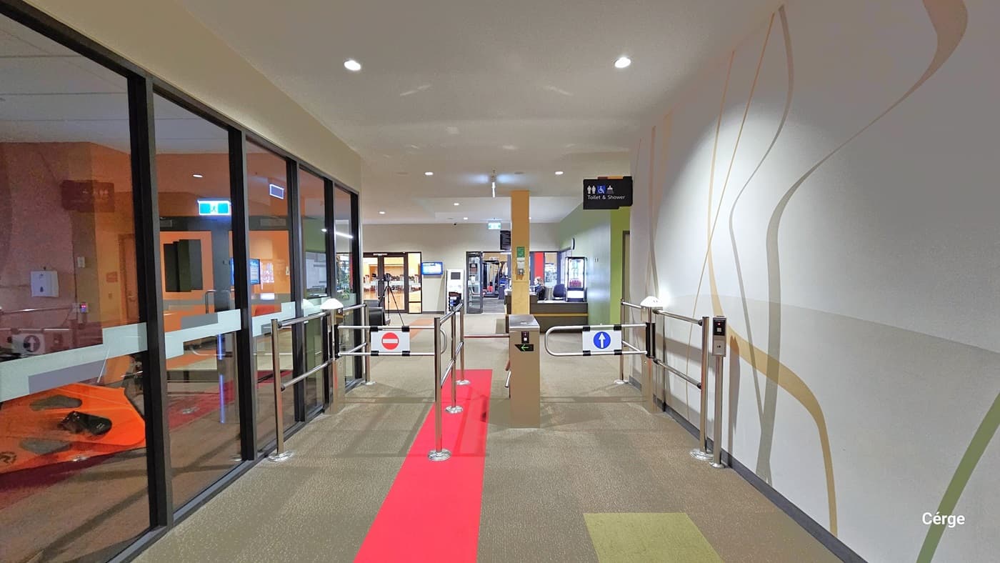 A corridor with a turnstile gate in the centre, featuring a red carpet leading through it. On the left, there is a glass wall with a view of an orange kayak inside a room. On the right, a white wall with abstract line designs. Above the gate, a sign indicates "Toilet & Shower" with directional symbols. The floor is carpeted in a neutral tone, and the ceiling is plain with recessed lighting.