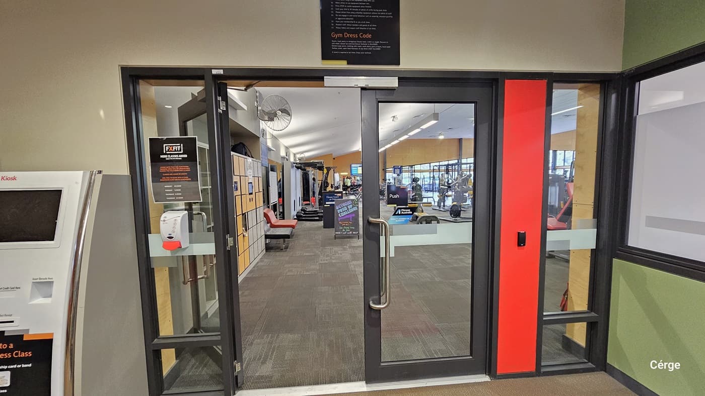Glass doors leading into a gym area. On the left, there's a kiosk with a screen. The doors have a sign with gym rules and a hand sanitiser dispenser. Inside, lockers line the left wall, and various gym equipment is visible further in. The floor is carpeted, and the ceiling has recessed lighting. A red vertical panel is on the right side of the door.