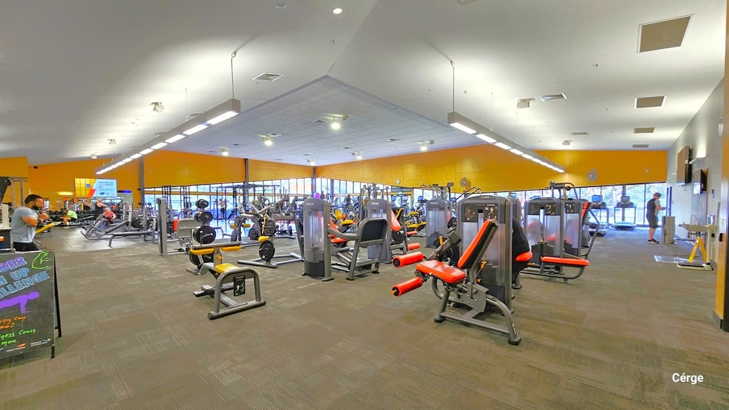 A spacious gym interior with various exercise machines arranged throughout. On the left, a person is using a machine, and a signboard is visible. In the centre, there are multiple weight machines with red and black padding. The right side shows more equipment and a person near the entrance. The floor is carpeted in a brown and green pattern, and the ceiling is fitted with fluorescent lights. Large windows in the background allow natural light to enter.
