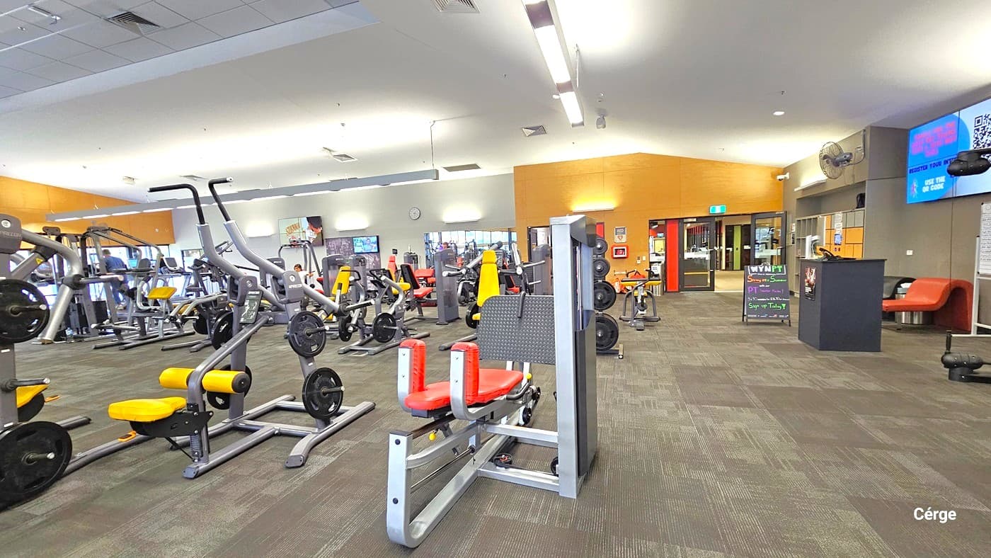 A spacious gym interior with various exercise machines on the left, including weightlifting and cardio equipment. In the middle, there are more machines and a few people working out. On the right, a reception area with a desk, a digital screen, and a seating area with an orange couch. The floor is carpeted in a green and brown pattern, and the ceiling is fitted with fluorescent lights. The walls are painted in orange and grey tones.
