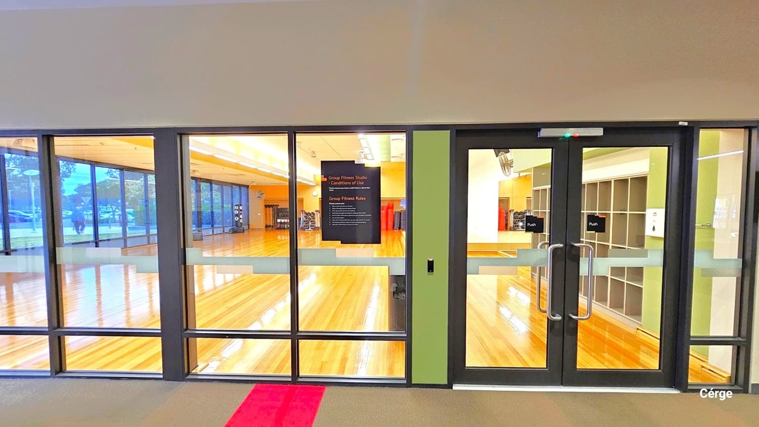 Glass doors with metal handles lead into a spacious room with a polished wooden floor. The room is well-lit with overhead lights and has large windows on the left side, allowing natural light to enter. Inside, there are exercise mats and equipment visible against the far wall. A sign is posted on the glass door. The ceiling is plain, and the floor outside the room is carpeted with a red strip running across it.