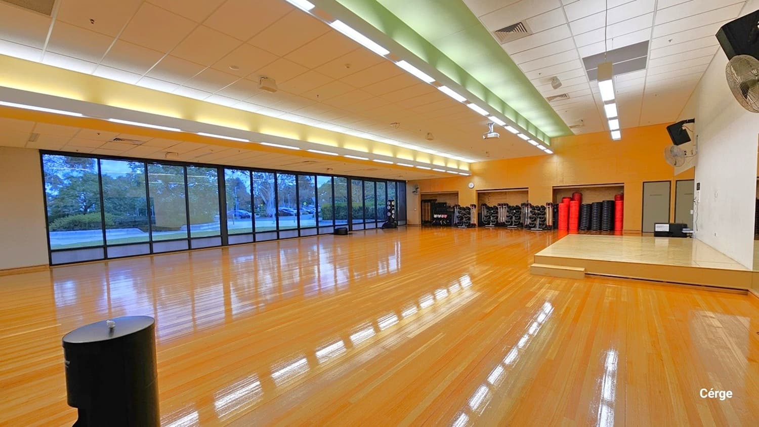 A spacious room with a polished wooden floor and a high ceiling featuring fluorescent lights. On the left, large floor-to-ceiling windows offer a view of trees and a road outside. On the right, a small stage is present with a fan mounted on the wall above. In the background, there are stacks of exercise mats and equipment against an orange wall. The room is well-lit and appears to be a fitness or multipurpose space.