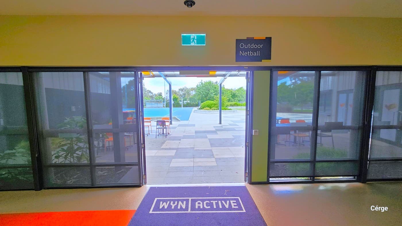 Entryway to the Outdoor Netball Courts at Eagle Stadium. Similar to other doors, this also has a black mat with WYN ACTIVE that is placed on the orange-painted section within the entrance doors. Above the door is a green sign for Exit and a black label for Outdoor Netball next to it. Outside is a seating area with orange and black plastic chairs.