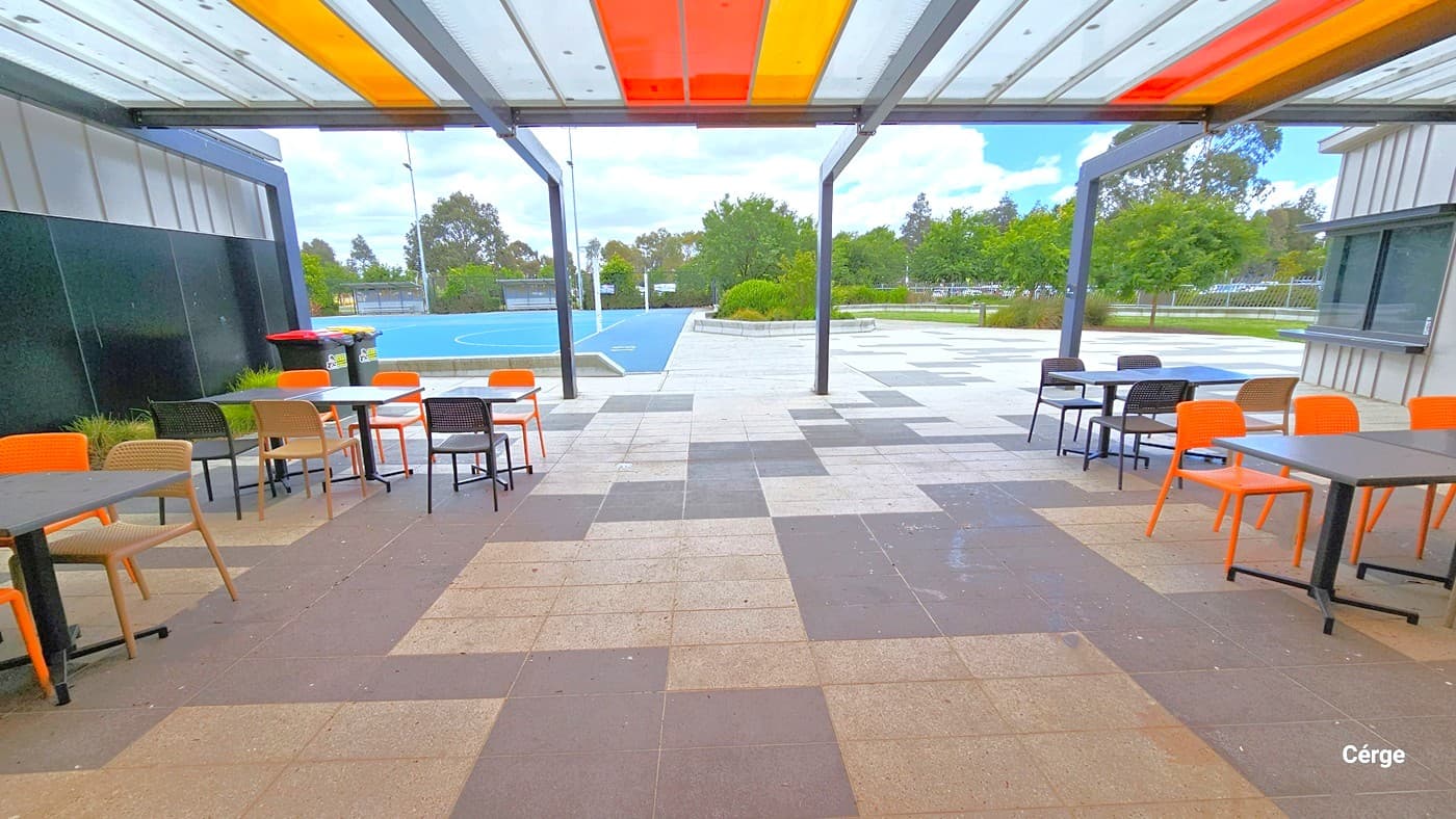 Seating of Outdoor Netball Courts is protected by a canopy cover made of colored polycarbonate sheets supported by metal beams. The outdoor seating has a grey square table top with black metal leg support and orange, brown, and black plastic chairs. The outdoor floor area is made of alternating tiles of dark grey and light grey tiles. Two large garbage bins with orange and yellow lids can be found near the left black side wall.