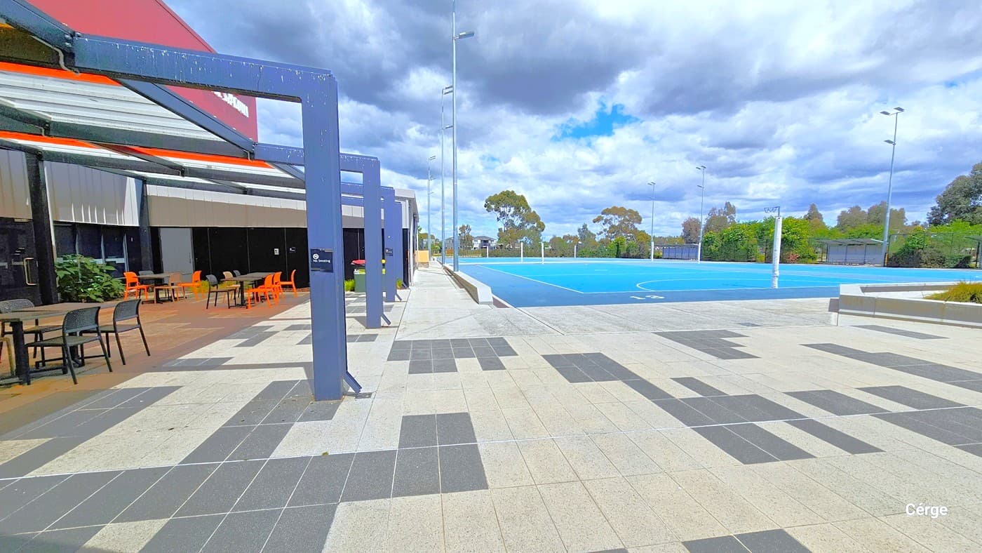 Image shows the outdoor seating and the Outdoor Netball court at Eagle Stadium. The outdoor floor area is made of alternating tiles of dark grey and light grey tiles. The outdoor seating has a grey square table top with black metal leg support and orange, brown, and black plastic chairs. On the large solid blue metal beams are black NO Smoking signs. Tall LED lights illuminate the court at night.