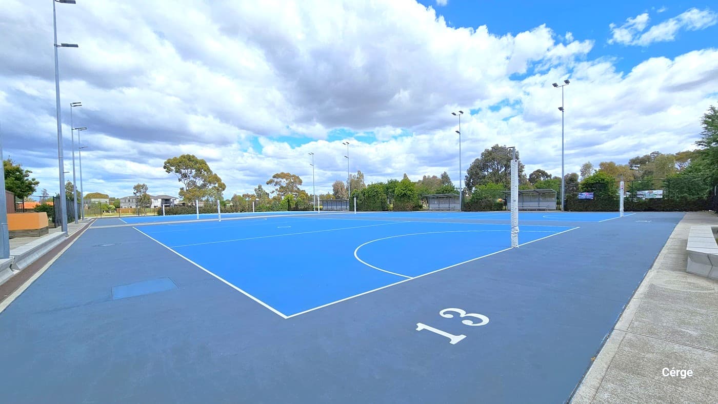 A closer look at the Outdoor Netball Court with the number 13 painted onn the purple-blue court surround. The Netball court has a blue floor with white lines for the goal circle, transverse line, and center circle. There are solid grey benches between the lamp posts and on other sides of the court. Further to the right side is a bench with shade. The property is enclosed by a black chain-linked fence.