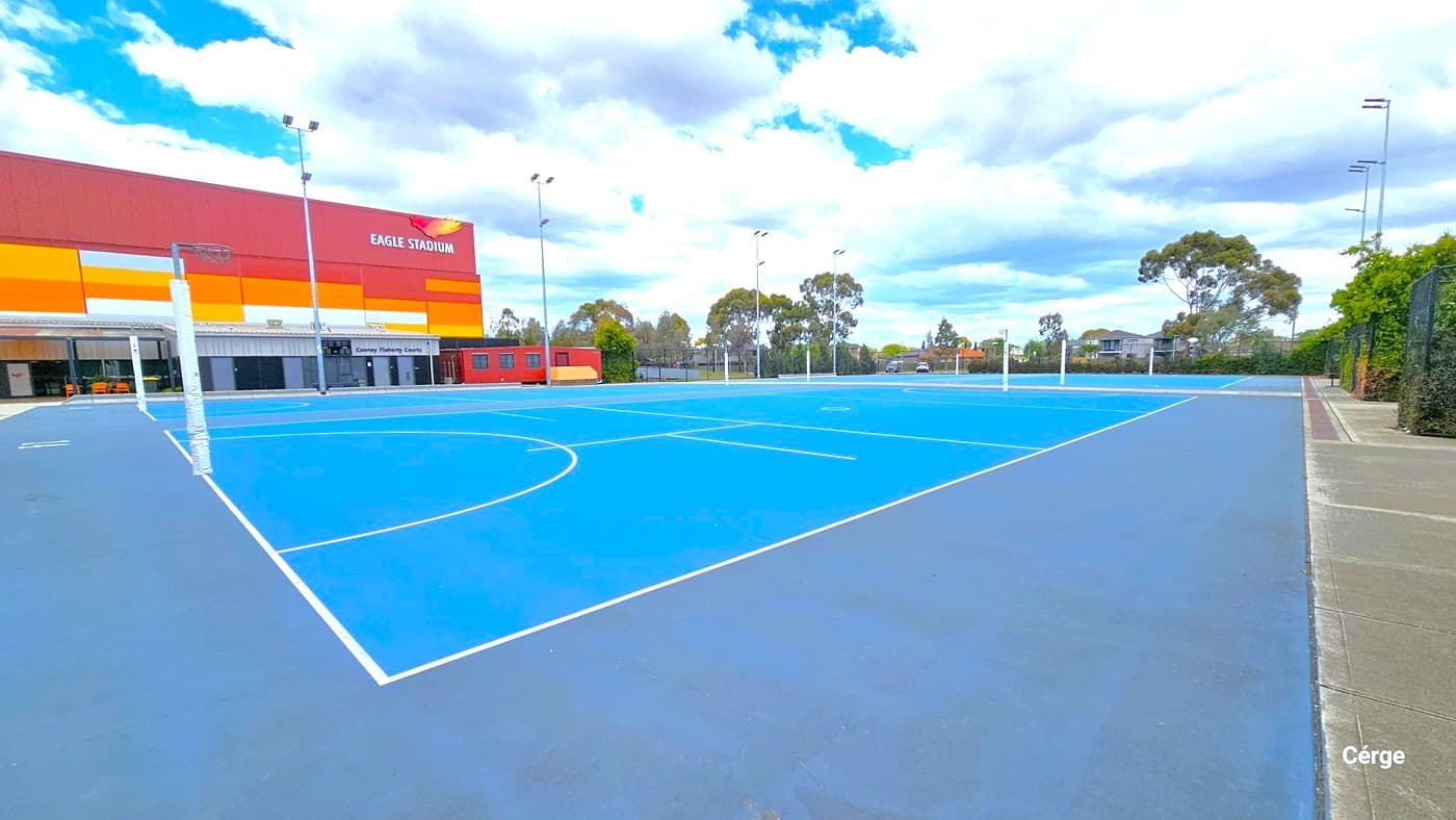 Another angle of the Outdoor Netball Court and the white goalpost placed within the white end line. The court has a blue floor with white lines and a purple-blue court surround. On the right end is a grey concrete pathway. Several tall lamp posts illuminate all the netball courts at night. An eagle logo above the Eagle Stadium is detailed on the red wall with yellow, orange, and white accents. Outside the fence are trees, vines, bushes, and residential buildings.
