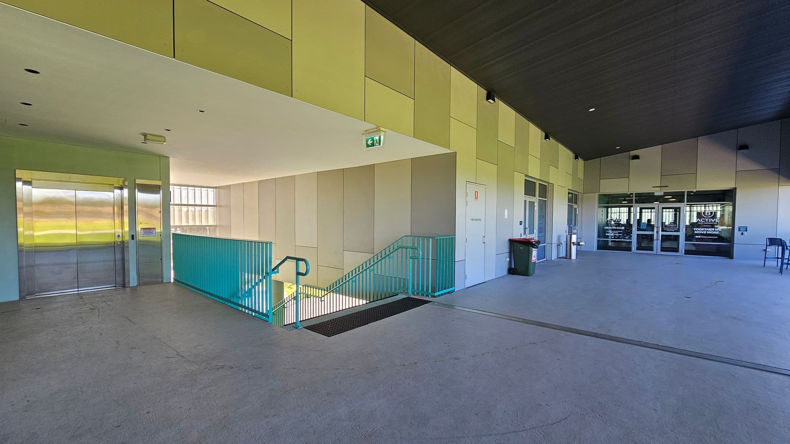A spacious indoor area with a grey floor and a ceiling featuring recessed lights. On the left, there is a lift with metallic doors and a green emergency exit sign above. A staircase with turquoise railings descends to a lower level. On the right, there are glass doors with signage, leading to another room. A green rubbish bin and a chair are positioned near the doors. The walls are covered with light-coloured panels, and a large window is visible in the background.