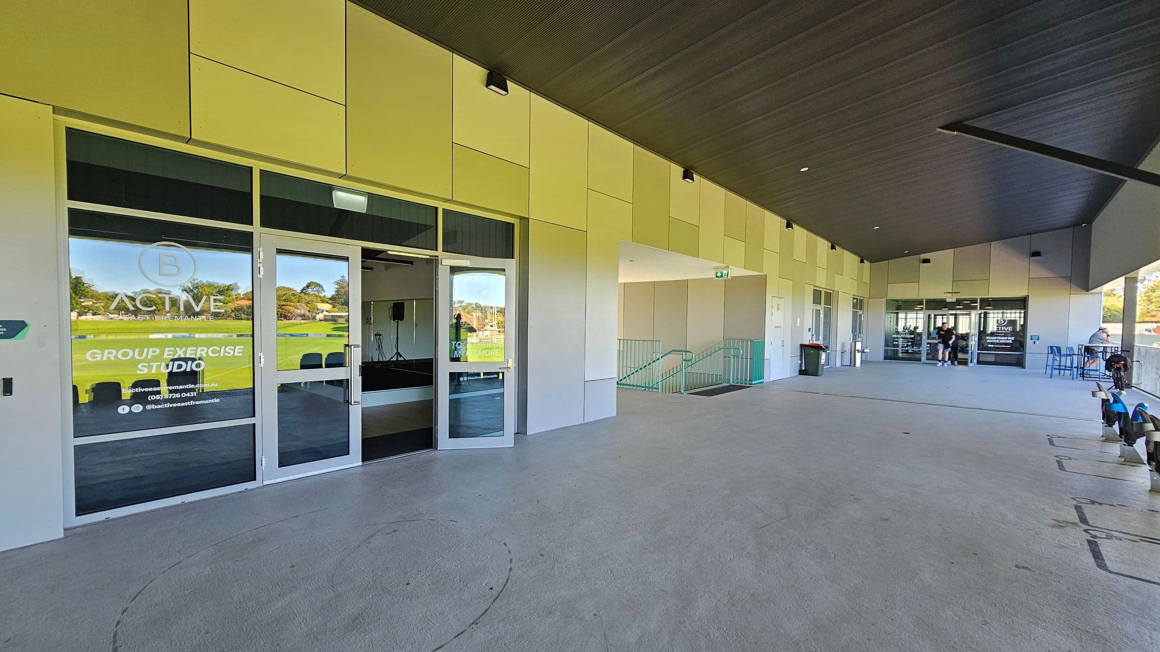 A covered outdoor area with a light-coloured tiled wall on the left featuring a glass door labelled "Group Exercise Studio." The floor is concrete, and the ceiling is dark with recessed lighting. In the middle, there is a small set of stairs with a green railing. On the right, there are glass doors leading to another part of the building, with a few people near them. Blue chairs and a bin are also visible on the right side.