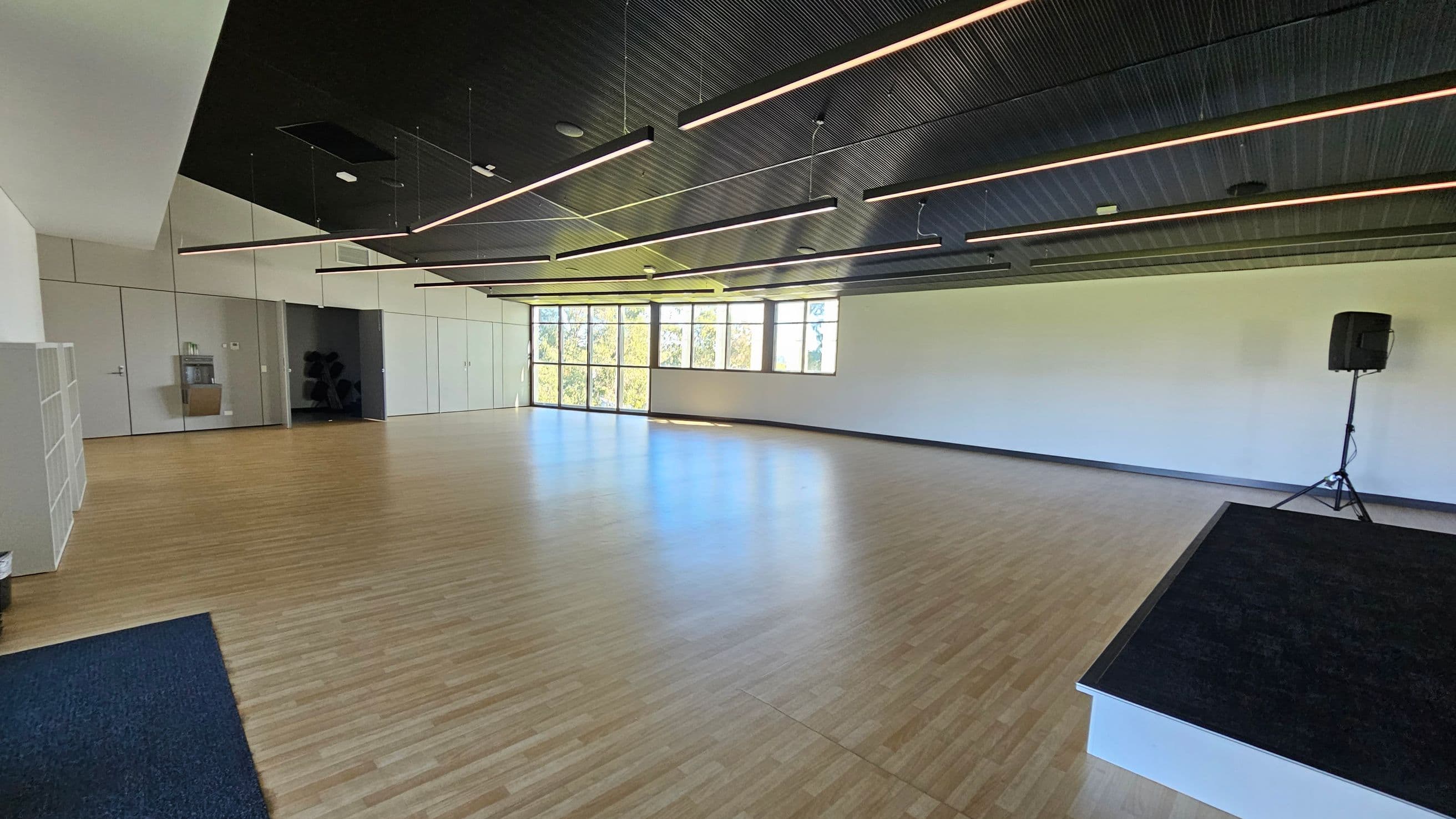 A spacious room with a wooden floor and a dark, ribbed ceiling featuring linear lights. On the left, there are white storage shelves and a door. The back wall has large windows allowing natural light to enter. On the right, a speaker on a stand is positioned near a small black platform. The room appears empty and well-lit.