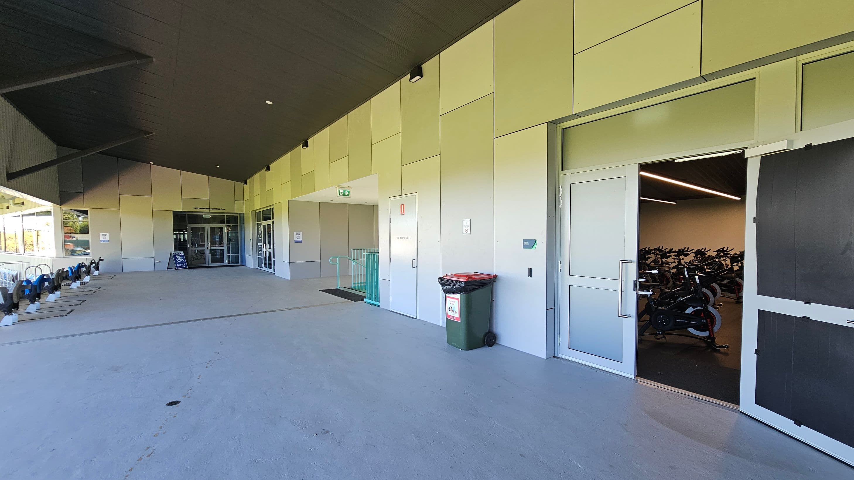 On the left, there are bicycle racks with several bikes parked. In the centre, a glass entrance door with a signboard in front leads into a building. To the right, a partially open door reveals a room filled with stationary exercise bikes. A green rubbish bin with a red lid is placed against the wall near the door. The ceiling is dark with recessed lighting, and the floor is smooth concrete.