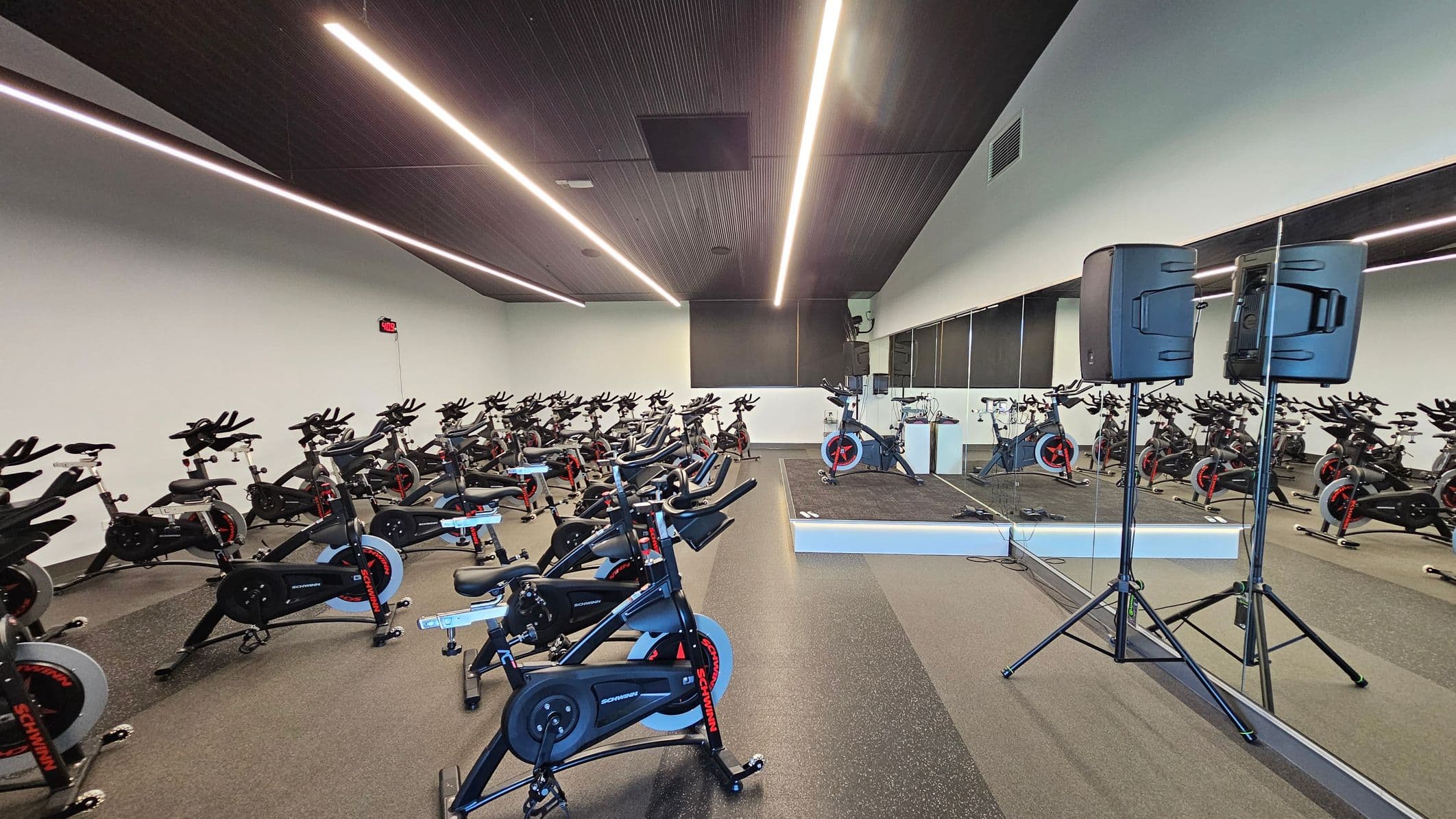 A spacious indoor cycling studio with rows of stationary bikes on the left and centre. The right side features a large mirror reflecting the bikes and a speaker on a stand. The floor is covered with dark, speckled rubber, and the ceiling has linear lights. A small stage with an instructor's bike is at the front. The walls are light-coloured, and a digital clock is mounted on the left wall.