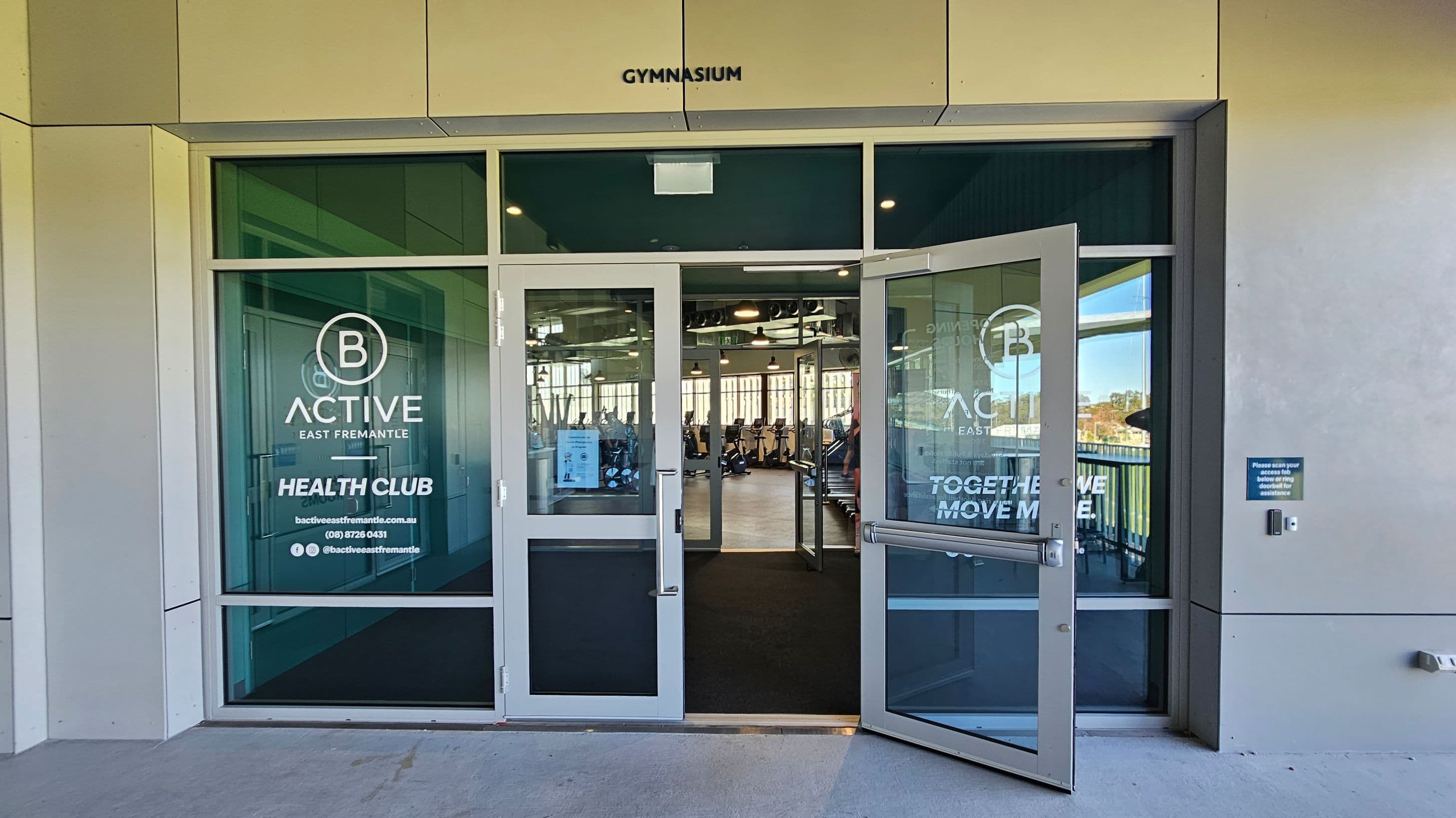 Entrance to a gymnasium with glass doors, one of which is open. The doors have signage for "B Active East Fremantle Health Club" with contact details. Inside, gym equipment is visible, including exercise bikes and treadmills. The exterior wall is light-coloured, and there's a sign on the right instructing access via a device. The floor is concrete outside and dark carpet inside. The ceiling inside has visible lighting and ventilation ducts.