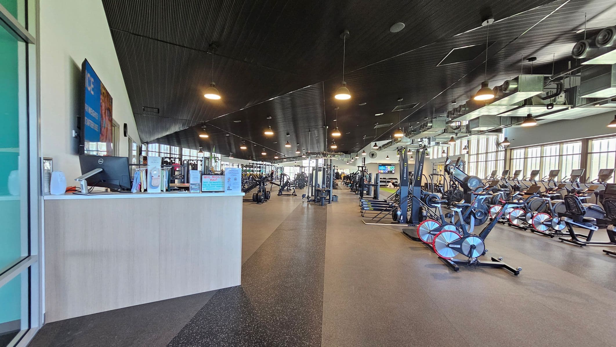 A spacious gym interior with a reception desk on the left, featuring a computer and informational materials. The middle area is filled with various exercise machines, including weightlifting equipment and stationary bikes. On the right, there are rows of treadmills and other cardio machines. The ceiling is dark with hanging lights, and large windows line the right wall, allowing natural light to illuminate the space. The floor is covered with a mix of rubber and carpeted surfaces.