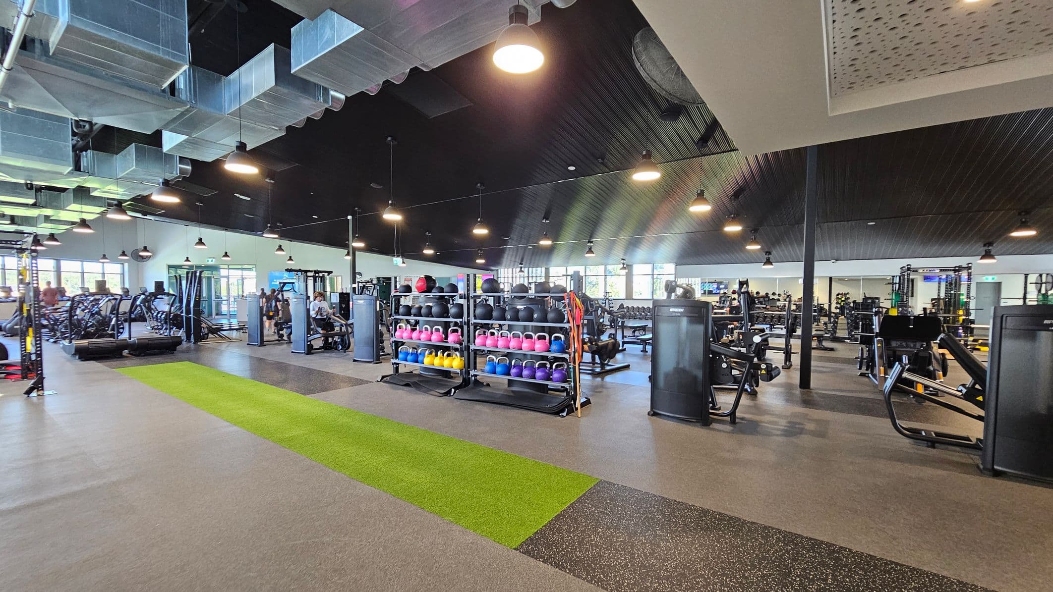 A spacious gym interior with a variety of fitness equipment. On the left, there are treadmills and elliptical machines near large windows. In the centre, racks of colourful kettlebells and medicine balls are neatly arranged. On the right, various weight machines and benches are visible. The floor is a mix of grey and green turf, and the ceiling features exposed ductwork and hanging lights. The gym is well-lit with natural and artificial light.