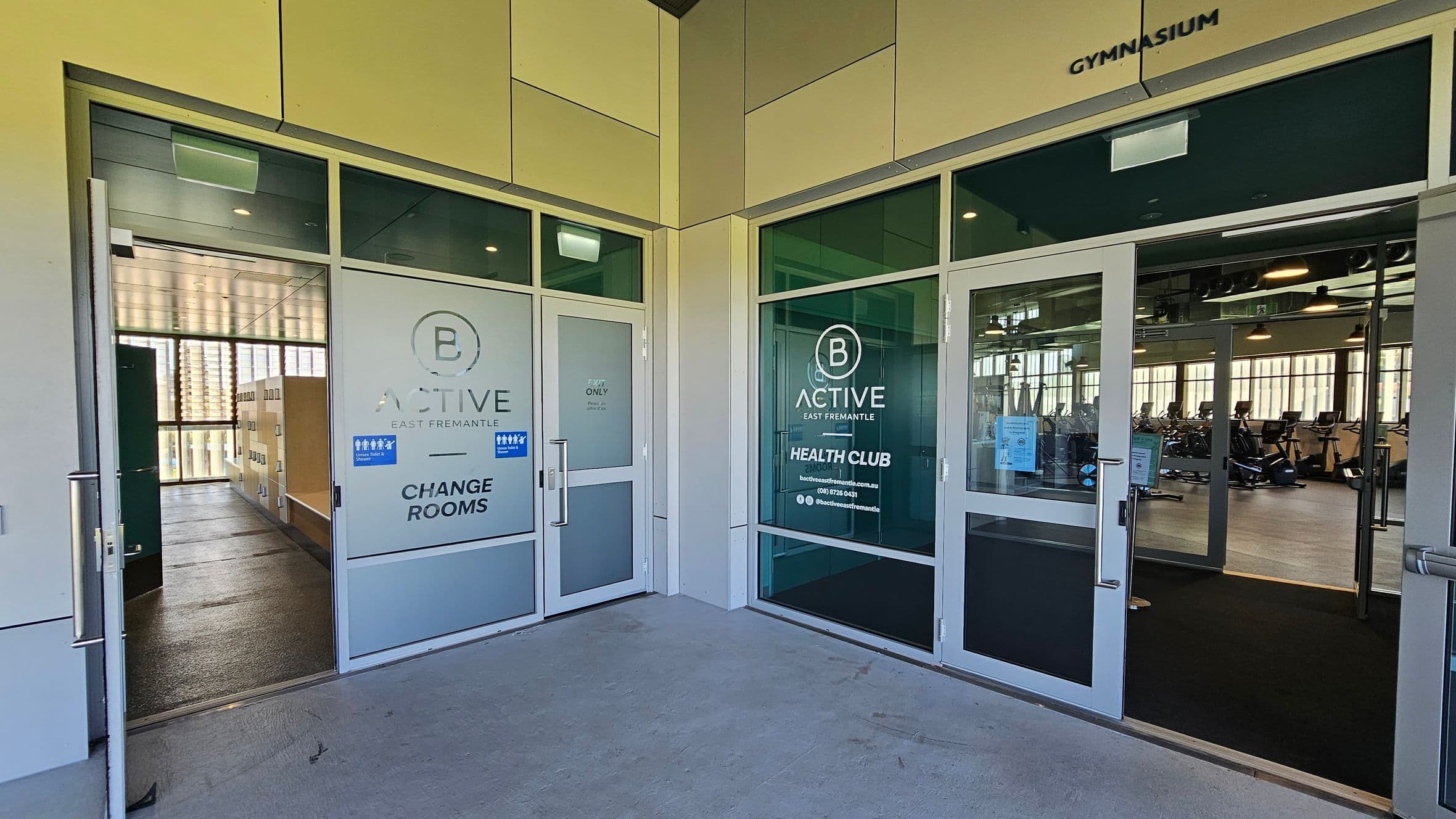 Entrance to a gymnasium with two glass doors. The left door leads to the change rooms, marked "B Active East Fremantle," with visible lockers inside. The right door leads to the health club area, also marked "B Active East Fremantle," showing exercise equipment like stationary bikes. The floor is concrete, and the ceiling has recessed lighting. The walls are a combination of glass and light-coloured panels.